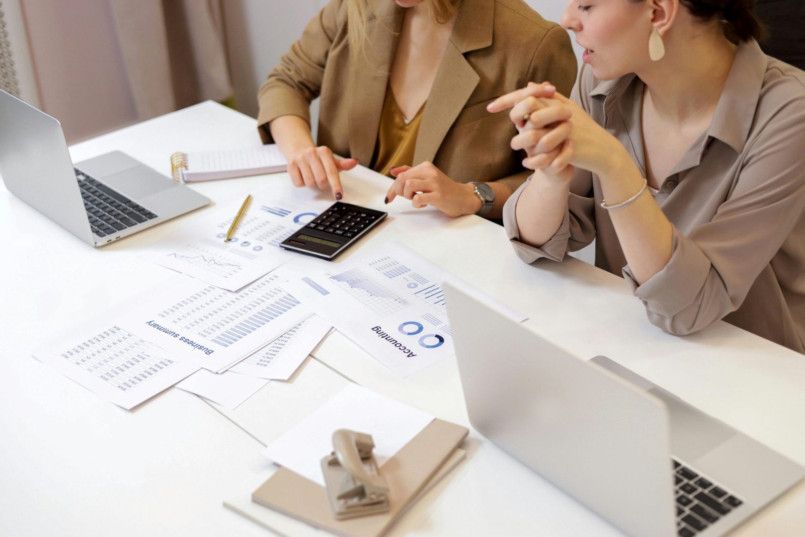 Two female colleagues collaborating on financial analysis at a modern office desk. They are reviewing "Business summary" and "Accounting" reports with graphs, using a calculator and laptops to discuss data.