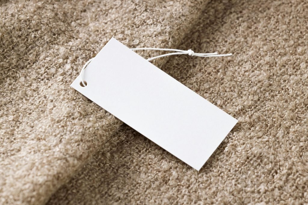 A blank white clothing hang tag with string resting on brown textured knitwear fabric.
