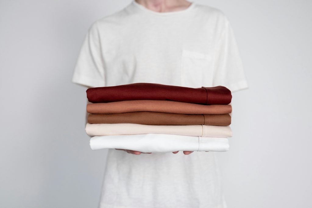 Man holding a stack of folded earth-tone cotton t-shirts in a minimalist studio setting.